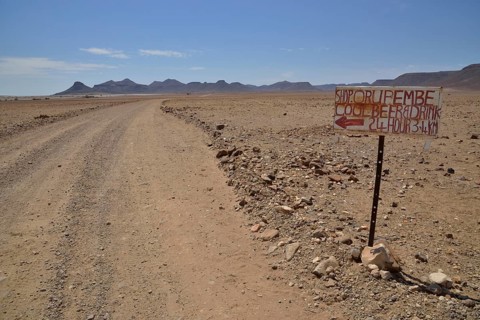 Hand-painted advertising sign for a liquor store on a dirt road in a barren landscape, Orupembe,
