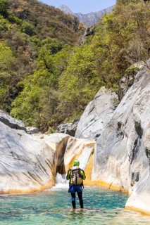 Back view of a canyoneer standing in a clear turquoise pool, preparing to climb a smooth natural