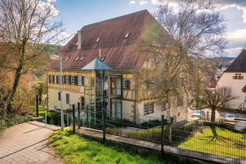 Timbered house with a large gable roof, surrounded by trees and a path in the foreground,