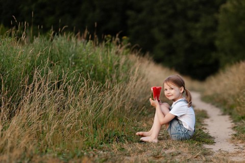 A child sits barefoot on a grassy path, enjoying a slice of juicy watermelon. The summer sun casts