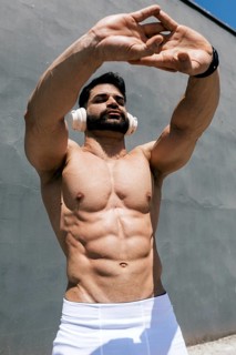 Muscular man exercising outdoors, wearing headphones, stretching under the sunlight. His toned