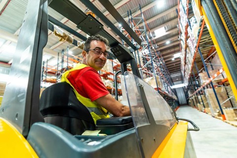 Happy manual worker turning to smile while driving forklift in distribution warehouse