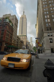 A classic yellow taxi is prominently featured in the foreground on a bustling Manhattan street,