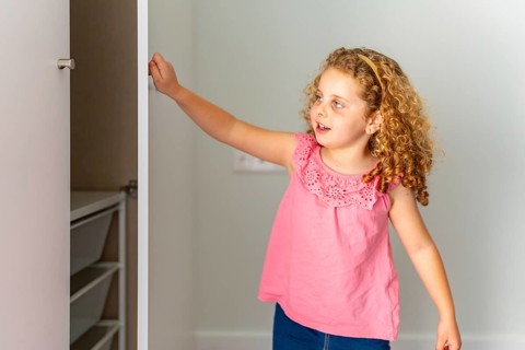 Young curly haired girl opens an empty wardrobe in her new room, exploring shelves and drawers with