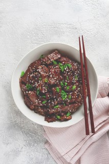 Crispy beef, with green onions and sesame seeds, in a bowl with chopsticks, on a textured surface,