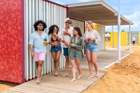Group of diverse friends walking barefoot on a wooden boardwalk, smiling and holding refreshing