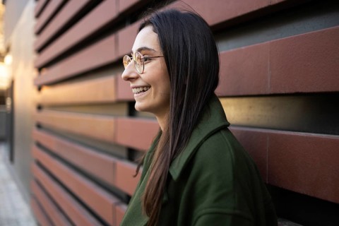 A woman wearing glasses and a green coat stands smiling in profile view against a textured wall The