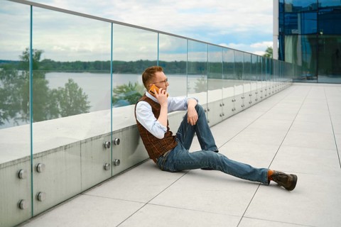 Middle-aged man sits on floor of observation desk beside body of water. He is making phone call