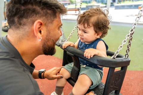 Father kneeling and comforting his sad toddler son sitting on a swing at the playground, creating a