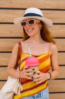 Woman smiling brightly wearing a sun hat and sunglasses, holding a colorful beverage with a straw