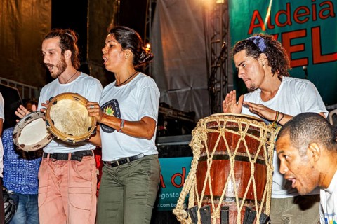 Three people passionately play drums and percussion instruments at a concert, a band plays and