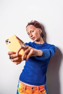 A trendy woman wearing colorful trousers and a blue top takes a selfie with her yellow phone,
