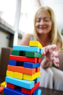 A smiling senior woman carefully removes a block from a colorful stacking tower. The bright colors
