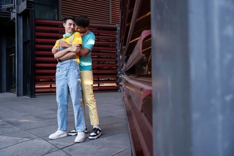 A gay couple share a tender embrace outside an urban building. They are dressed casually, exuding