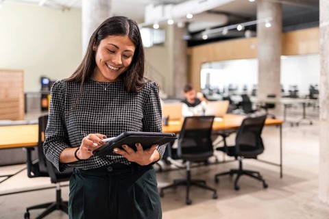 A woman in a modern office smiles as she uses a tablet, with colleagues in the background The image
