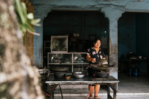 A woman in a floral dress cooks delicious traditional food in an outdoor market stall. The scene