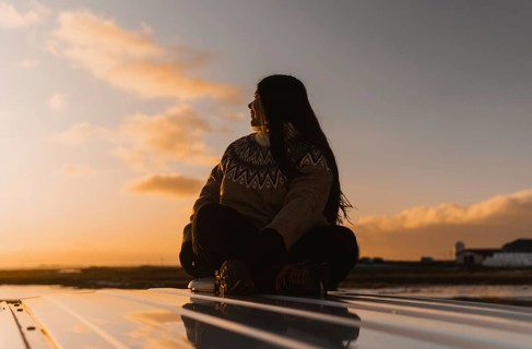 Colombian woman sitting calmly on a camper roof, gazing towards a picturesque Iceland sunset,