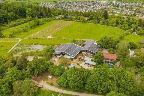 A farm with solar roofs surrounded by green fields and trees near a village, Sportplatz Aidlingen,