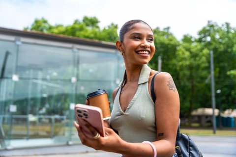 Young woman smiling while holding a coffee cup and using a smartphone, enjoying a casual moment in