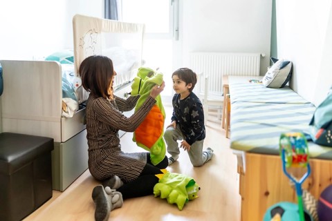 Mother holding a dinosaur costume while her son is kneeling on the floor in his bedroom, ready to