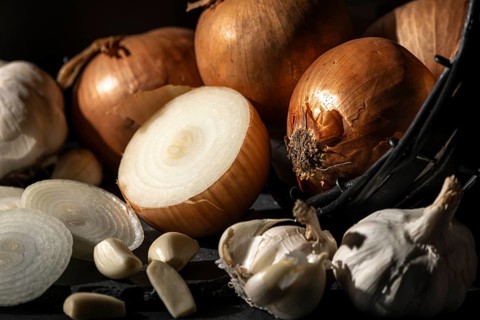 Whole and sliced onions and garlic next to a basket, warmly lit