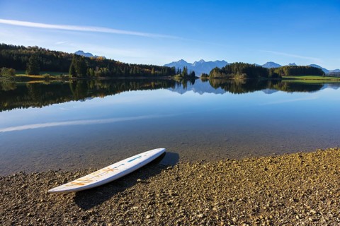 A calm lake with a surfboard on the shore, surrounded by mountains and a clear blue sky, Forggensee