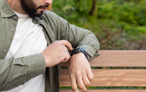 A man interacts with his smartwatch while sitting on a wooden bench in a lush in the city. He wears
