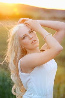A close-up portrait of a blonde woman in a white dress against a sunset in a grassy field