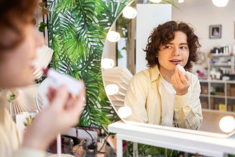 A young man applies lipstick while looking into a mirror surrounded by lights in an organic beauty