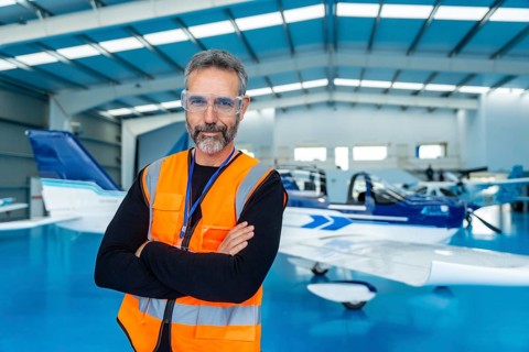Mature bearded aviation technician in orange safety vest and protective glasses stands with arms