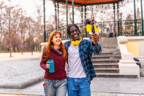 Two young content creators are recording a vlog in a park, holding a smartphone with a microphone