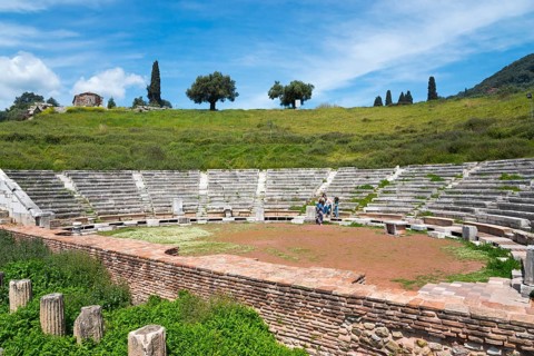 An ancient amphitheatre with surrounding green landscape and hills under a partly cloudy sky,