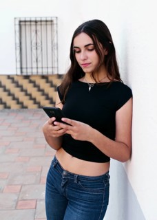 A young woman in casual jeans and a black top leans against a white wall, focused on her smartphone