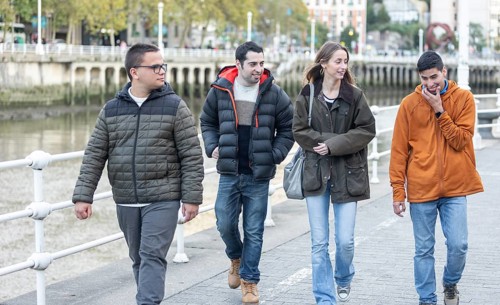 Caregiver walks alongside young men with intellectual disabilities along a riverside promenade.