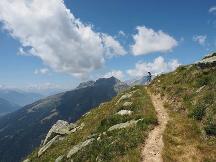 Female hiker on a trail through mountainous terrain under blue sky with clouds, Bellwald, Goms,