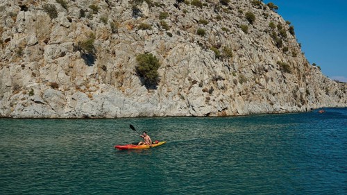 A person paddles in a kayak along steep rocks in blue water, Rina, harbour, Vathi, Vathys,