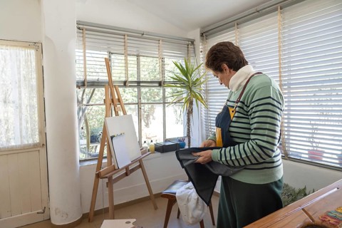 Older woman in striped sweater examines or folds a dark denim apron while standing near a wooden