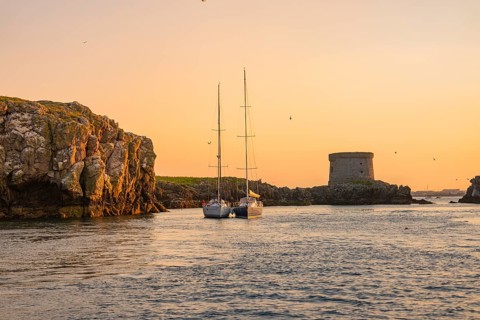 A sailboat sails between rocks near a historic tower at sunset, Howth, Ireland