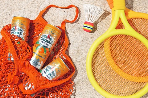 In a mesh bag, on a sandy beach, there are cans of tangerine lemonade and Borjomi drink. There are