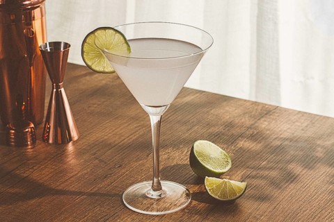 A clear cocktail sits in a glass with a lime slice on the rim. A copper shaker and halved lime are