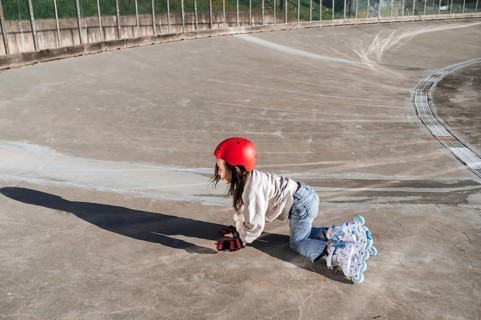 A young girl with a red helmet and protective gloves cautiously gets up from the ground, from a