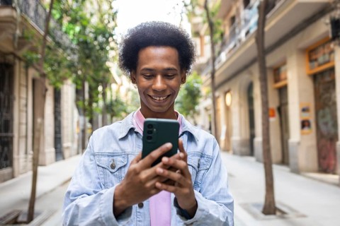 A non-binary person with an afro hairstyle smiling while using a smartphone in a vibrant urban