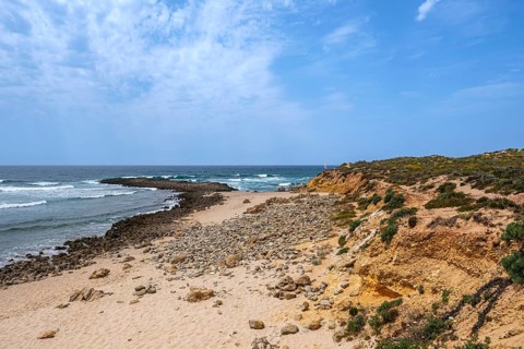 Quiet beach with rocks surrounded by blue skies and gentle ocean waves, Vila Nova de Milfontes,