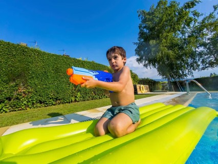 Young boy on a green inflatable float in a sunny pool, squirting a water toy and laughing as he
