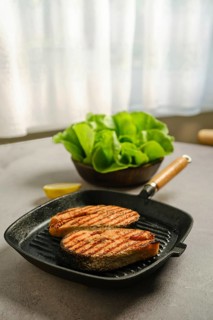 Grilled salmon steaks on frying pan on kitchen tabletop. Bowl of fresh salad sits on the table.