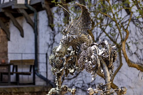 Metal sculpture of a dragon with detailed wings in front of a wall covered with vines, The fountain