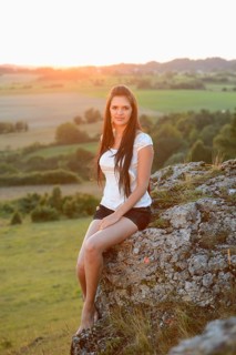 A woman with long hair sits on a rock in nature during sunset, wearing a white shirt, Bavaria