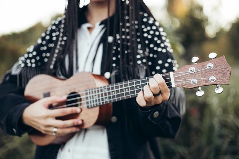 A woman in a black jacket plays a wooden ukulele against a blurred backdrop of grass during sunset,