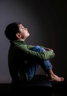 Young boy sits on a stool in a dark room, looking upward thoughtfully. His green shirt and jeans