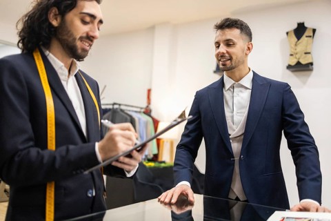 A tailor consults with a client in a suit store, taking notes while discussing suit options. They